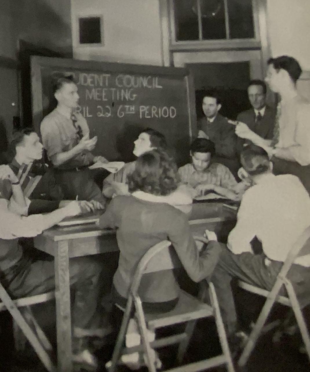 students sit around a desk and talk. Behind them, a chalkboard reads: student council meeting 6th peroid.